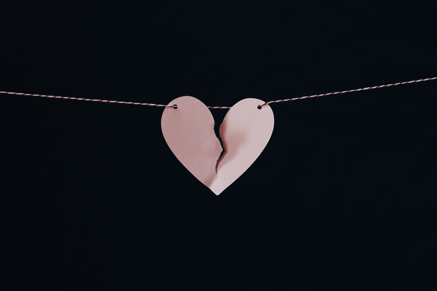A pink paper heart is hangs from a string across a black background. The heart is ripped nearly in two.