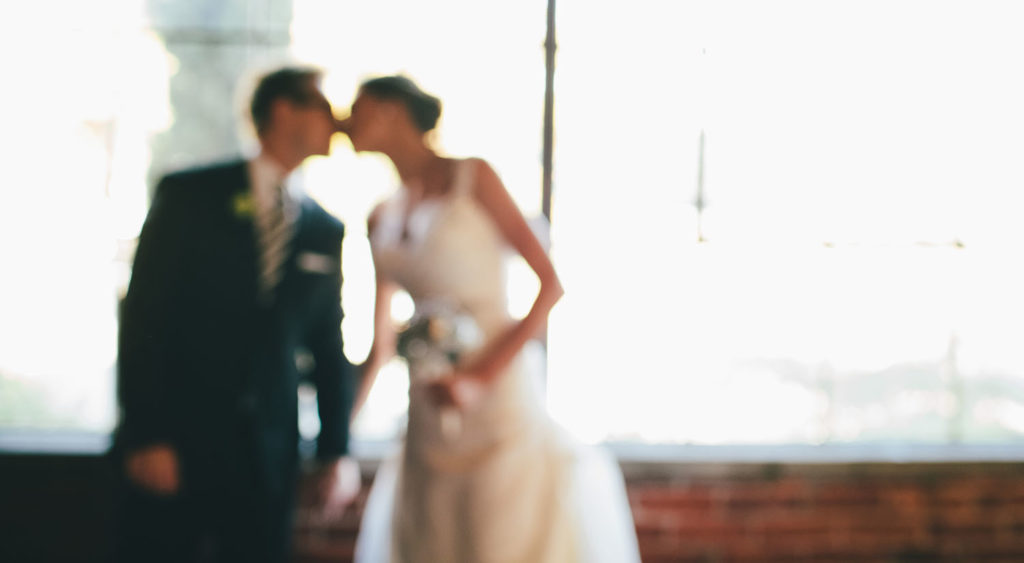 A blurred photo of a wedding couple kissing in front of a window. By Anne Simone