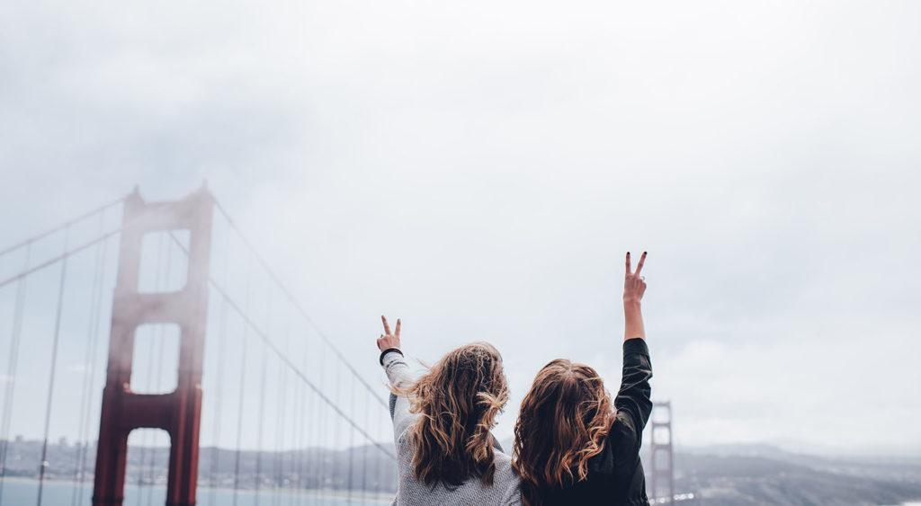Ian Schneider's Unsplash photo of two people with long hair with their backs to the camera while they make peace signs overlooking the Golden Gate Bridge in San Francisco